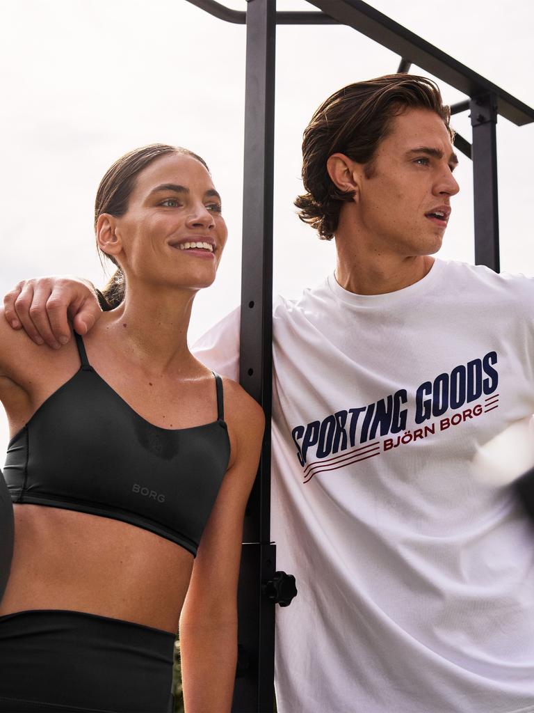 Woman in a sports bra and man in a "Sporting Goods" t-shirt stand together outdoors, smiling, near exercise equipment.