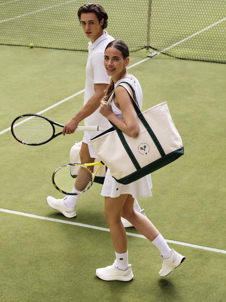 Two people in white tennis outfits walk on a grass court. One carries a large tote bag with a heart emblem. Both hold tennis rackets.