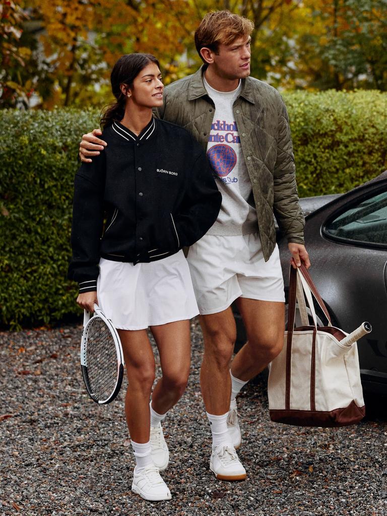A man and woman in casual tennis attire walk together, carrying a bag and tennis racket, surrounded by greenery and a parked car.