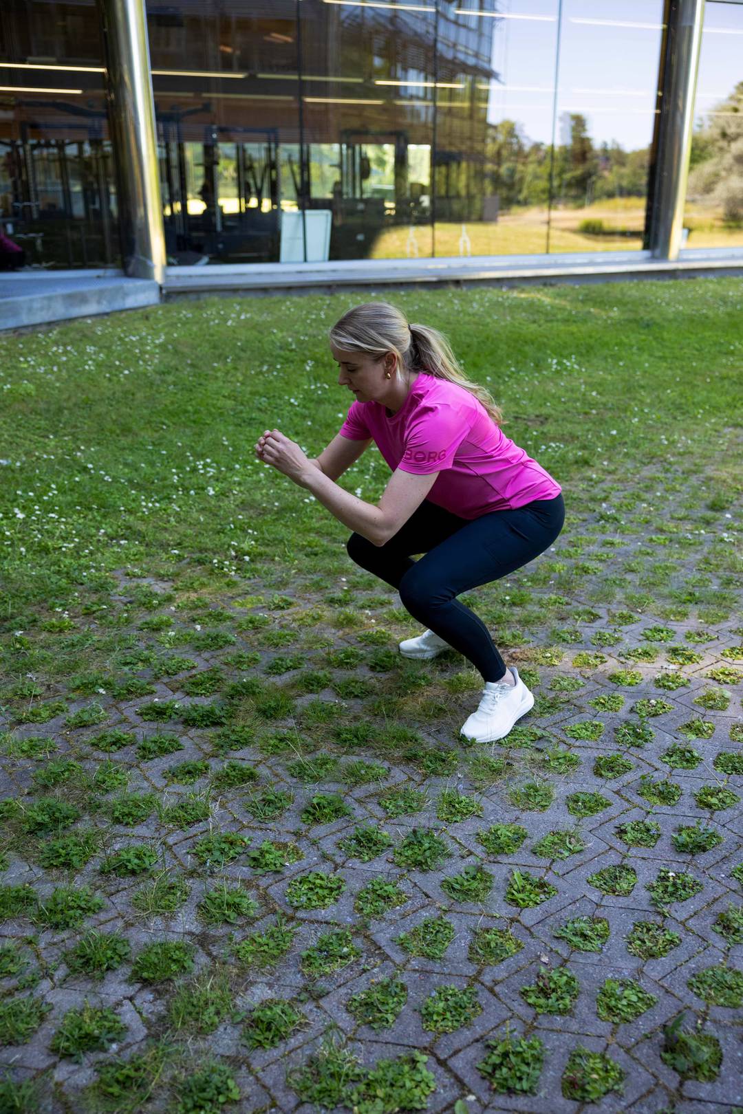 A blond woman outdoors in a pink Borg T-shirt, black Borg Running Tights and white Björn Borg sneakers, doing a jump squat