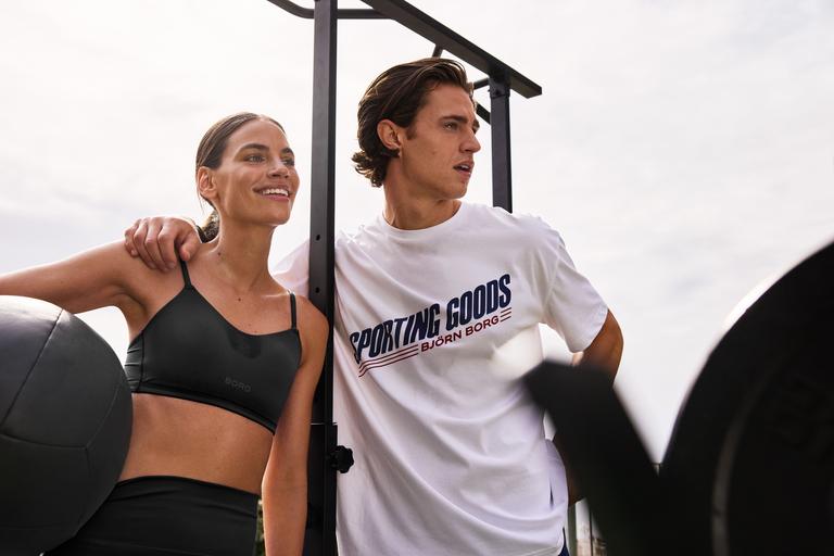 A man and woman in athletic wear stand together outdoors, with the man holding a medicine ball. Both appear focused and engaged.